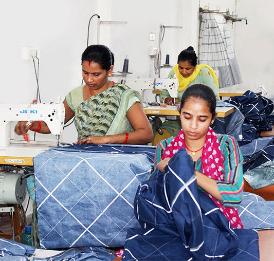 Three women sewing blue textiles on machines in a workshop setting.